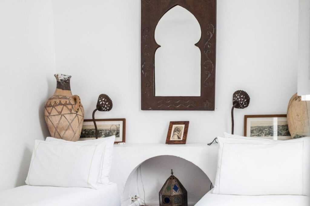 9 UNIQUE Riads in Tangier 17 Two white twin beds with white pillows sit against a white wall, reflecting the simple and rustic charm found in riads in Tangier. Between them are clay vases, framed photos, and a lantern. Above, a decorative arched mirror hangs on the wall.
