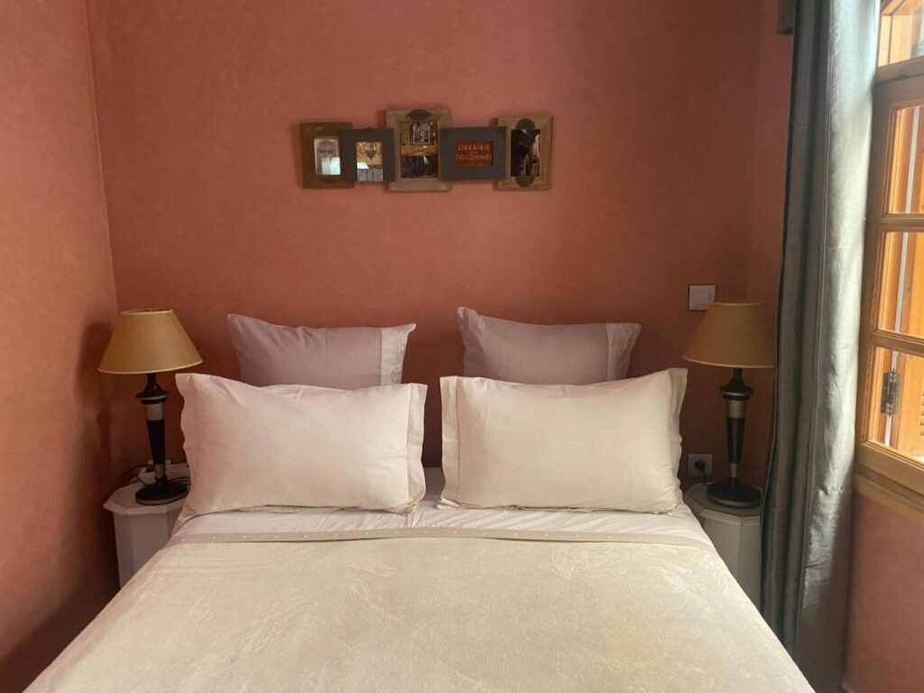 9 UNIQUE Riads in Tangier 34 A neatly made bed with white bedding and pillows is flanked by two bedside tables, each with a lamp. Reminiscent of elegant riads in Tangier, the room has peach-colored walls, framed art above the bed, and a window letting in natural light.