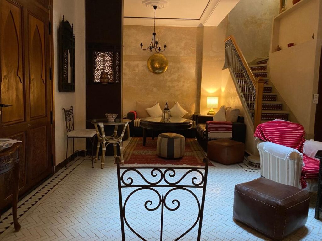 9 UNIQUE Riads in Tangier 37 A cozy living room inspired by riads in Tangier, featuring a patterned tile floor, a round black coffee table, sofas with cushions, a red striped throw, a staircase with decorative tiles, and warm lighting from lamps and a chandelier.