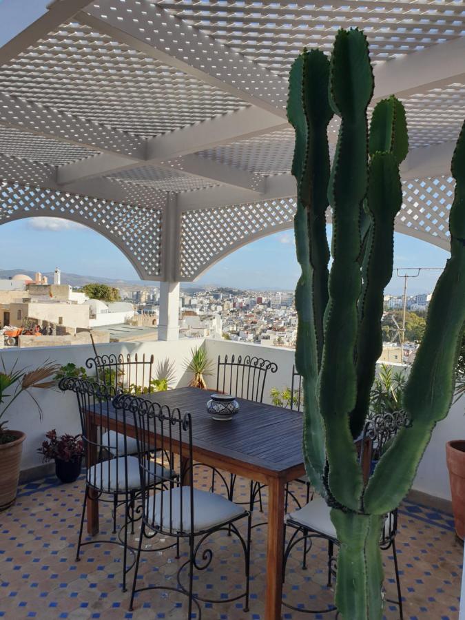 9 UNIQUE Riads in Tangier 36 A rooftop patio reminiscent of riads in Tangier features a wooden dining table, black metal chairs with white cushions, potted plants, and a large cactus. A lattice pergola offers shade, while the cityscape and blue sky create a picturesque backdrop.