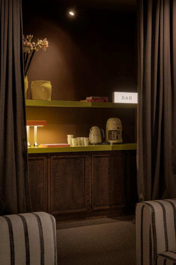 Where to Stay in Madrid: 14 Best Hotels 6 A cozy home bar setup—perfect for planning where to stay in Madrid first time—features a coffee maker, glassware, books, and a glowing BAR sign on green shelves, framed by dark curtains. Striped armchairs invite you to relax in style.