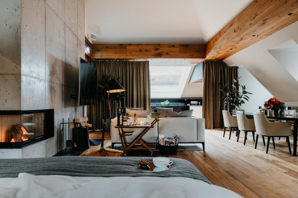 Authentic Boutique Hotels in Munich 23 A cozy, modern living space inspired by boutique hotels in Munich, with wooden floors, exposed beams, a lit fireplace, sofa, dining area, indoor plants, large windows with brown curtains, and a tea set on the table in the foreground.