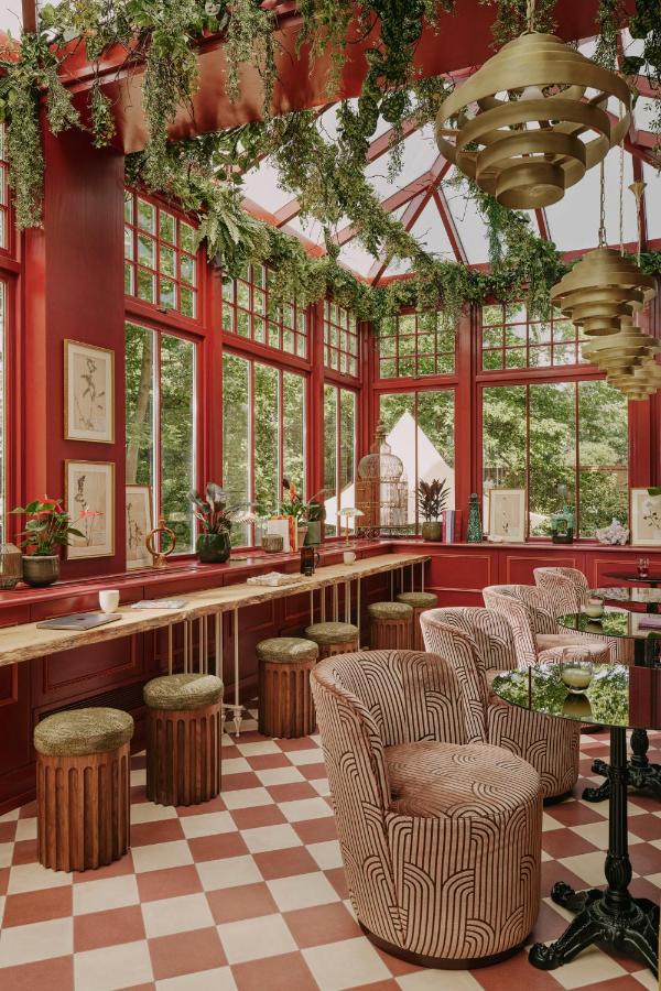 15 Best Boutique Hotels in London, UK 5 A sunlit room with red walls and large windows, reminiscent of the best boutique hotels in London, features hanging plants, patterned chairs, wooden stools, a marble countertop, and checkerboard tiles. Art and potted plants enhance the inviting space.