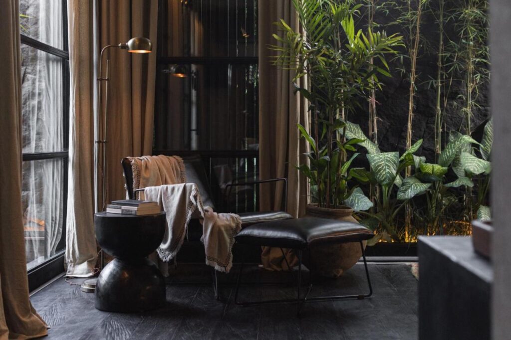 14 UNIQUE Boutique Hotels in Canggu, Bali 5 A cozy reading nook with a black leather chair, matching ottoman, blanket, small side table with books, floor lamp, and lush green plants by a large window with beige curtains—echoing the relaxing vibe of boutique hotels in Canggu.