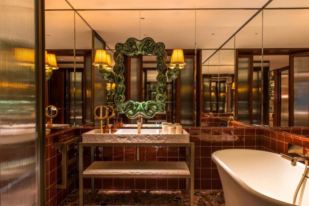 Where to Stay in Madrid: 14 Best Hotels 26 Luxurious bathroom with a large ornate green mirror, marble sink, brown tiled walls, and gold fixtures—perfect inspiration for those searching where to stay in Madrid first time. Mirrored walls enhance the elegant decor and freestanding bathtub.
