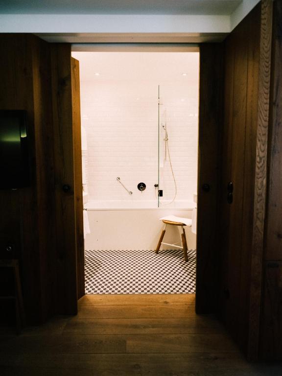 Unique Boutique Hotels in Salzburg 38 A bathroom in the style of boutique hotels in Salzburg, featuring white tiled walls and a black-and-white checkered floor, seen through open wooden doors. There’s a bathtub with a glass shower screen and a small wooden stool beside it.