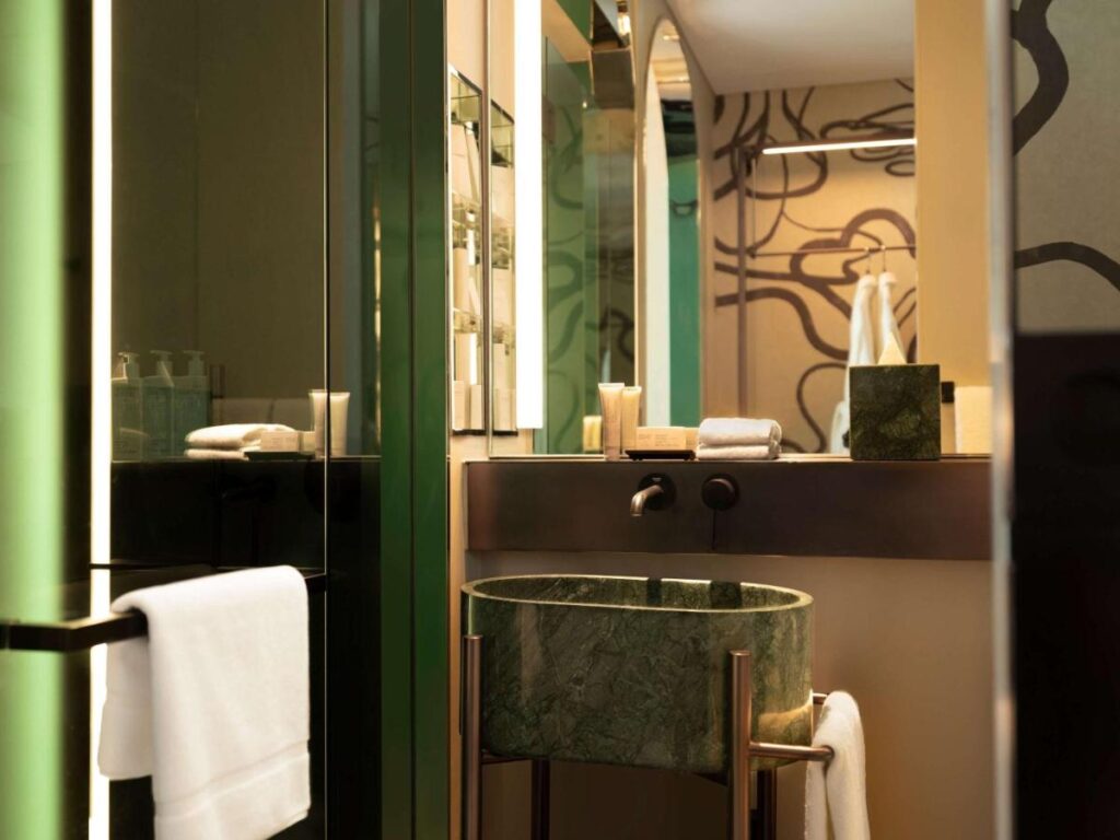 13 Most Unique Hotels in Singapore for an Incredible Holiday 51 Modern bathroom featuring a green marble sink, wall-mounted faucet, towels, and toiletries, with a mirror and abstract wall art—reflecting the sleek, contemporary design found in unique hotels in Singapore with warm lighting.