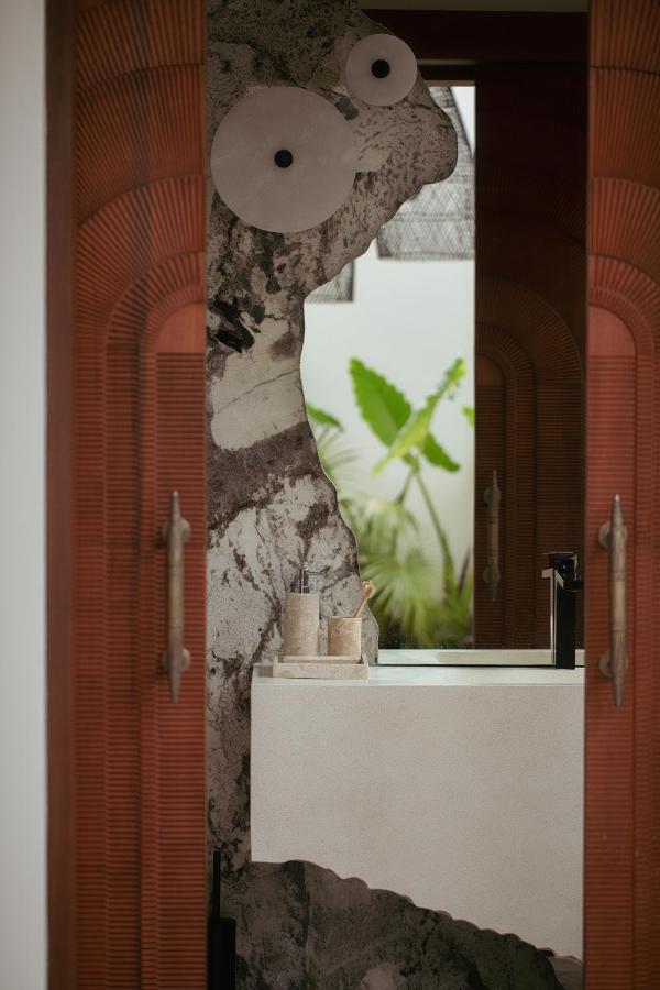 14 UNIQUE Boutique Hotels in Canggu, Bali 39 A bathroom with a modern sink and two containers, featuring a wall with an abstract design resembling large cartoon eyes—like artful touches found in boutique hotels in Canggu. A leafy plant is visible through a window in the background.