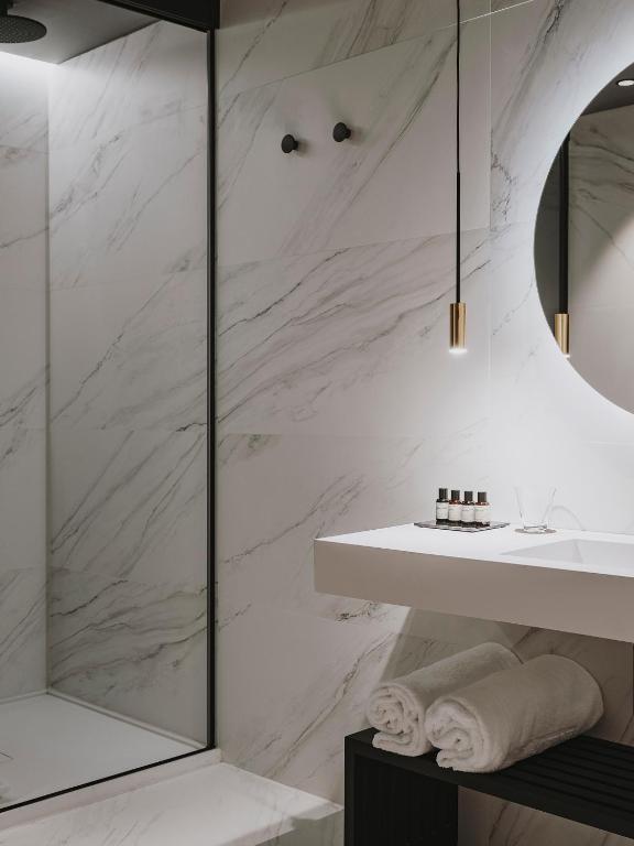 A modern bathroom inspired by boutique hotels in Asturias, with white marble walls, a glass shower enclosure, a round mirror, a floating sink with toiletries, and two rolled white towels on a black shelf underneath.