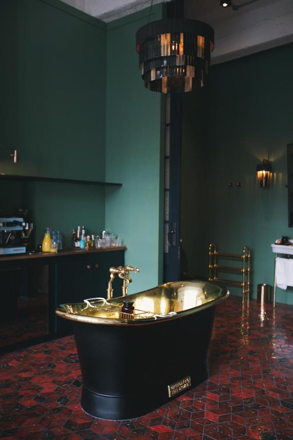 11 Unique Hotels in Georgia You Can't Miss! 3 A stylish bathroom in one of the unique hotels in Georgia, featuring a shiny brass tub, dark green walls, a modern black and gold chandelier, red tiled floor, and a countertop with drinks and glassware in the background.