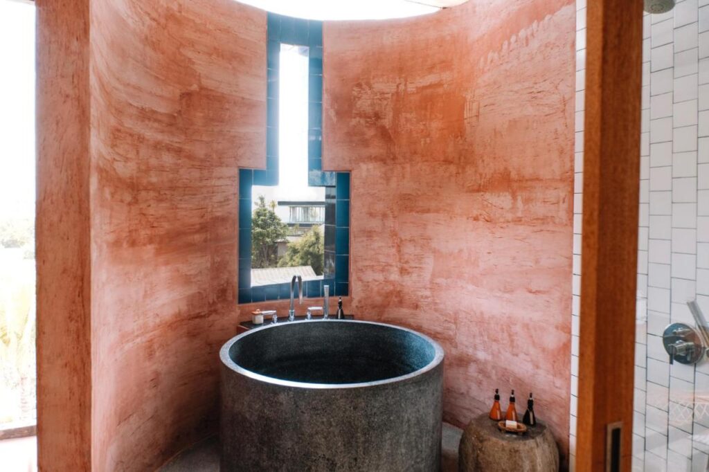 14 UNIQUE Boutique Hotels in Canggu, Bali 20 A modern bathroom inspired by boutique hotels in Canggu, with curved pink textured walls, a round stone tub, blue tile accents, and bath products on a wooden stool. Natural light fills the serene space.