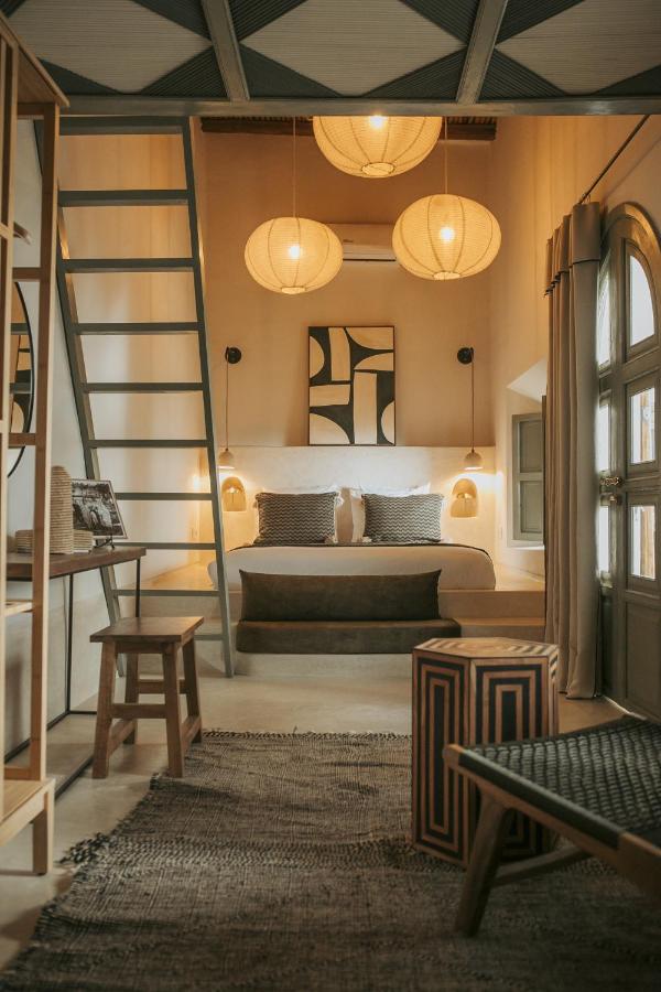 12 Charming Budget Riads in Marrakech 9 A cozy, modern bedroom inspired by budget riads in Marrakech, featuring a loft ladder, wooden furniture, soft lantern lighting, a double bed with cushions, abstract wall art, and large curtained windows in warm, earthy tones.
