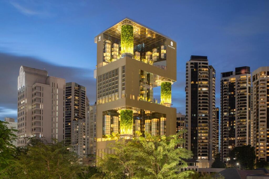 13 Most Unique Hotels in Singapore for an Incredible Holiday 43 A modern, multi-level skyscraper with illuminated green vertical gardens stands among other high-rise buildings at dusk, showcasing one of the unique hotels in Singapore, with trees in the foreground and a twilight sky in the background.