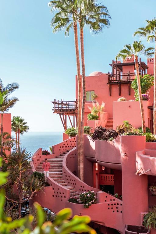 11 UNIQUE Boutique Hotels in Tenerife 46 Curved pink building with terraces and stairways, surrounded by tall palm trees and lush greenery, overlooking the ocean under a clear blue sky—one of the most stunning boutique hotels in Tenerife.