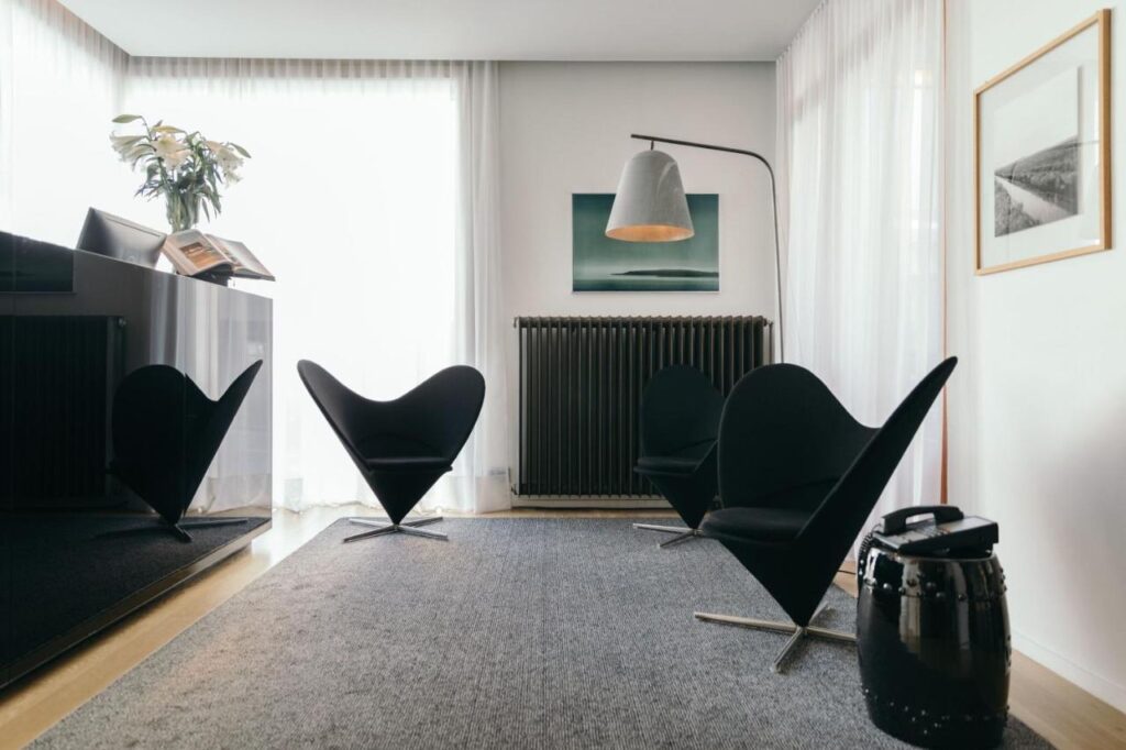 Where to Stay in Reykjavík: Top Guide 8 A modern living room, perfect for those seeking where to stay in Reykjavik, features three black heart-shaped chairs, a floor lamp, gray rug, wall art, light through sheer curtains, and a large plant atop a side table near the radiator.