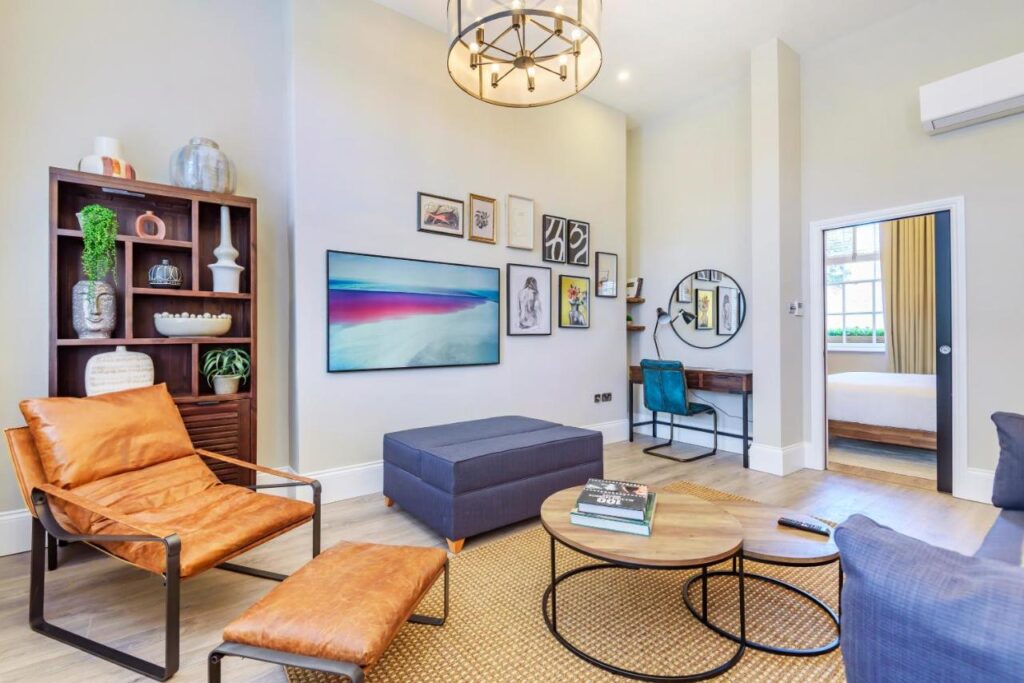 15 Best Boutique Hotels in London, UK 24 Modern living room with tan leather chairs, a blue ottoman, round coffee tables, a wall-mounted TV, bookshelf, desk, and decorative art on the walls—styled like the best boutique hotels London offers. A doorway leads to a bright bedroom.