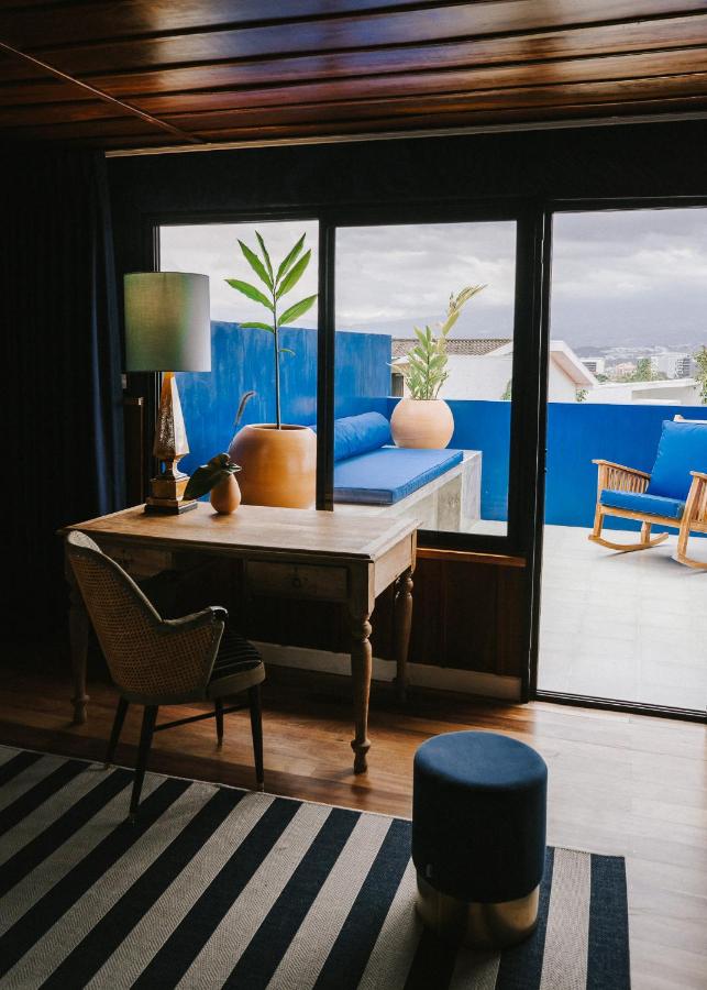 Unique Hotels in Costa Rica: Top Stays 16 A cozy room with a desk, chair, and lamp beside large windows. Outside, a terrace features blue walls, potted plants, and a lounge bench—reflecting the charm found in unique hotels in Costa Rica under a cloudy sky.
