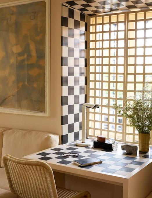 25 Weekend Trips from Madrid Max. 2h Away 61 A cozy workspace with a checkered black-and-white desk and matching wall pattern by a window with a grid screen—perfect for planning weekend trips from Madrid. A lamp, vase with flowers, notebook, glasses, and small decor items adorn the desk.