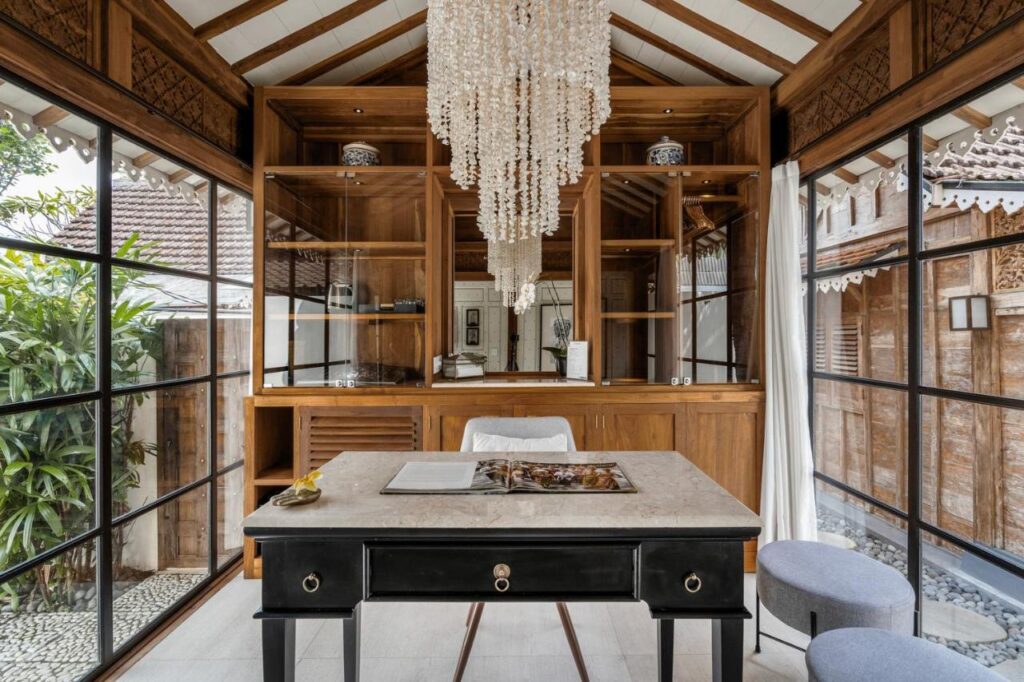 14 UNIQUE Boutique Hotels in Canggu, Bali 43 A stylish home office with glass walls, a black desk with an open book, a chandelier, and wooden shelves channels the cozy, natural feel of boutique hotels in Canggu, with lush greenery outside and ample natural light streaming in.