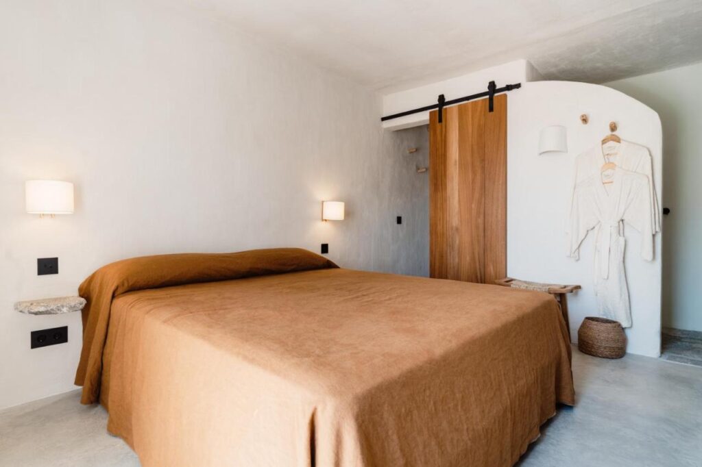10 AUTHENTIC Finca Hotels in Ibiza 20 Minimalist bedroom with a large bed covered in a brown blanket, two wall-mounted lamps, a wooden sliding door, and hanging bathrobes—echoing the serene style found in finca hotels in Ibiza. A wicker basket rests on light-colored floors and walls.