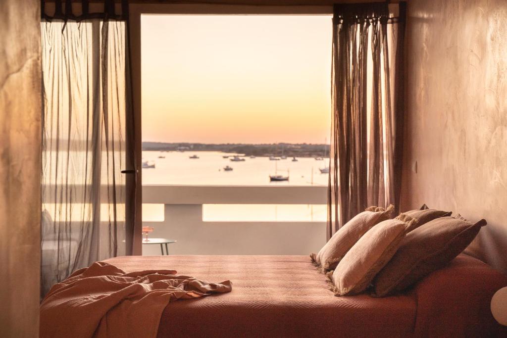 A cozy bedroom with a neatly made bed is bathed in warm sunset light, reminiscent of boutique hotels in Formentera. Sheer curtains frame a sliding glass door, which opens to a balcony overlooking a calm bay filled with boats.