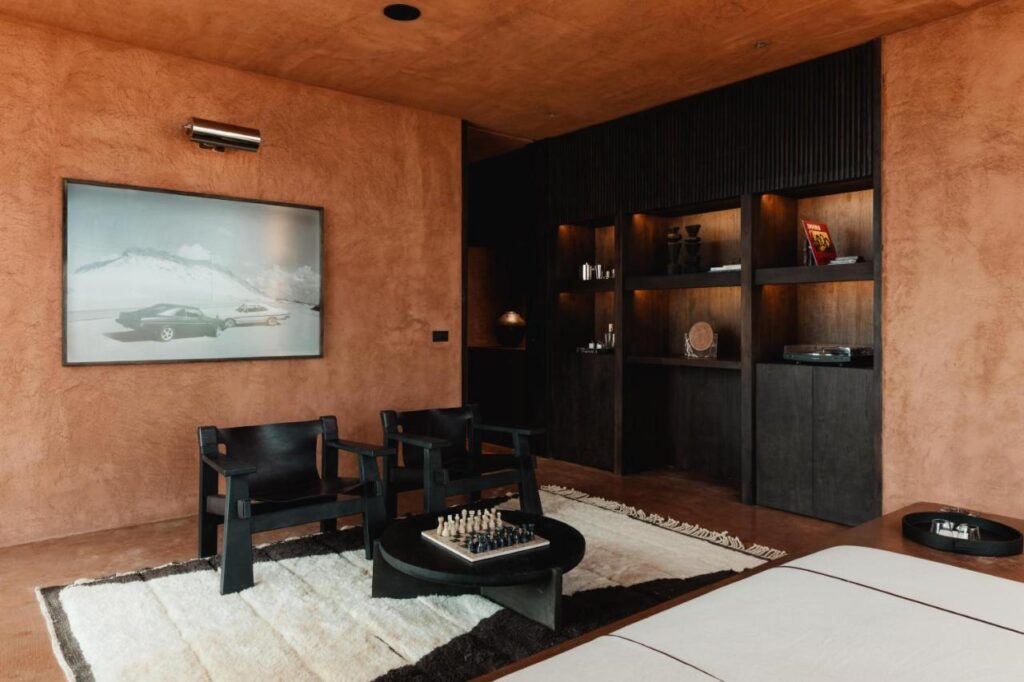14 UNIQUE Boutique Hotels in Canggu, Bali 12 A modern room with textured brown walls, two black chairs, a round coffee table with a chess set, a large framed car photo, an open dark wood shelving unit with books and decor, and a white and brown rug—reminiscent of boutique hotels in Canggu.