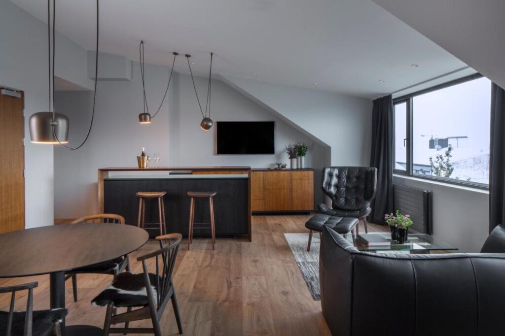 Where to Stay in Reykjavík: Top Guide 14 Modern loft-style living room with wooden floors, black leather sofa, lounge chair, and wall-mounted TV—ideal for those wondering where to stay in Reykjavik. Enjoy a kitchen bar with stools and a snowy view featuring a ski lift outside the large window.
