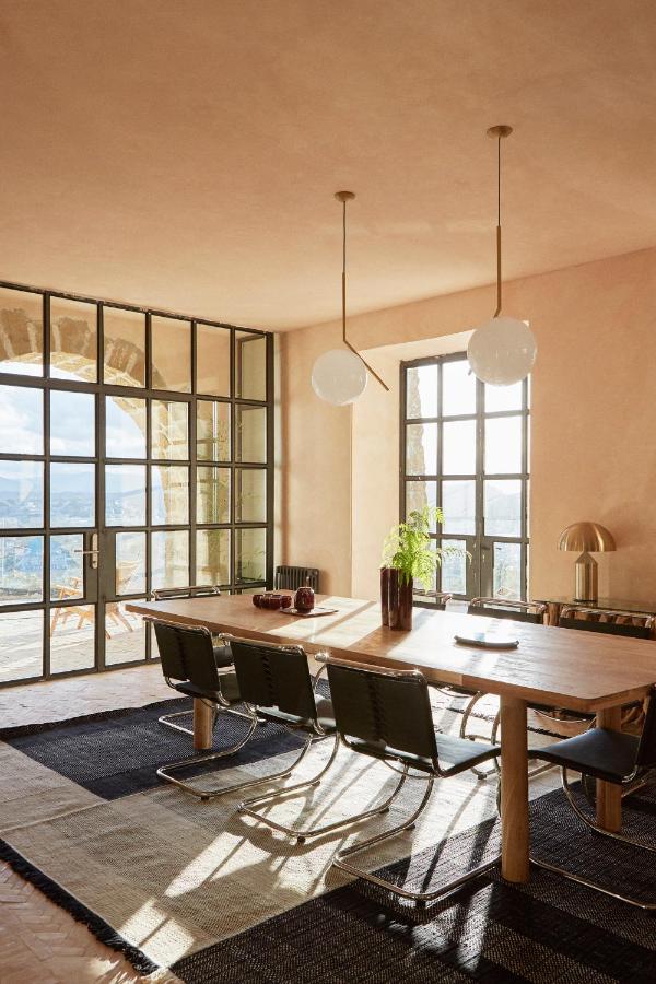 Best Boutique Hotels in San Sebastian 22 A sunlit dining room with a large wooden table, six black chairs, modern pendant lights, and large windows offering an outdoors view—reminiscent of the stylish interiors found in boutique hotels in San Sebastian. Plants, vases, and a gold lamp add chic touches.