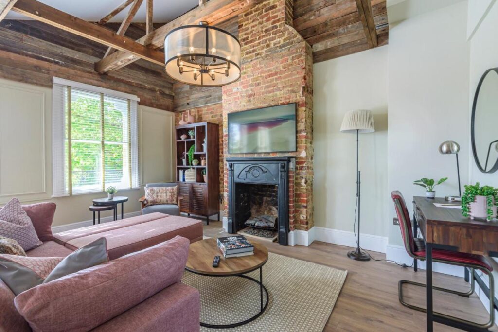 15 Best Boutique Hotels in London, UK 23 A cozy living room with exposed brick and wood beams evokes the charm of the best boutique hotels London offers, featuring a pink sectional sofa, round coffee table, fireplace with TV above, small desk area, and sunlight streaming through the window.