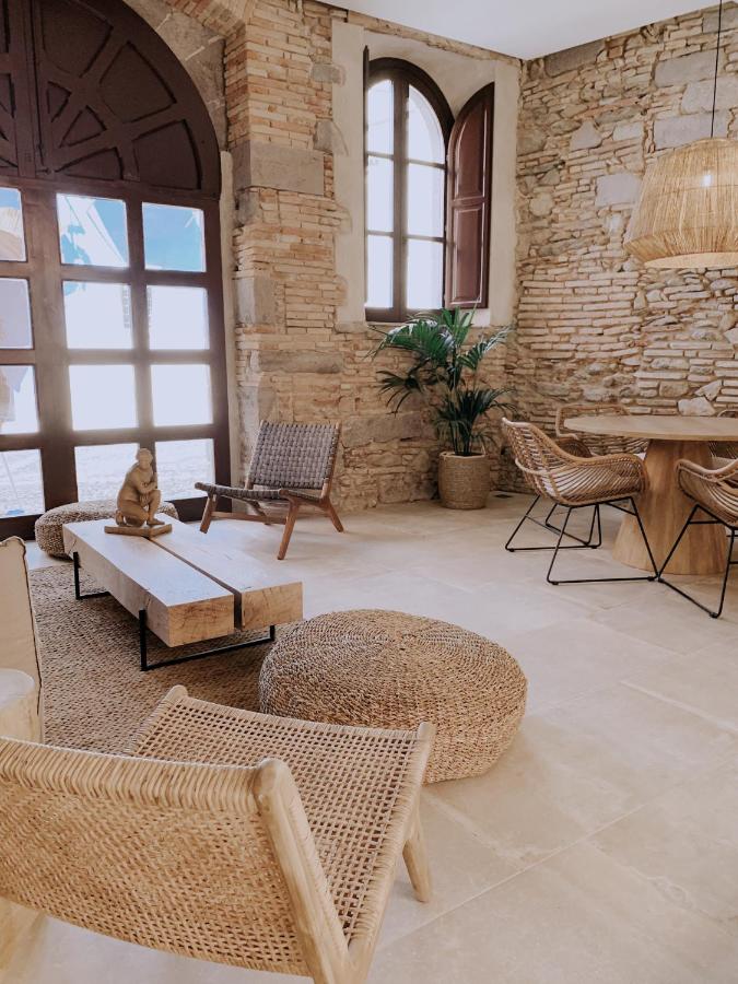 A rustic living room reminiscent of boutique hotels in Costa Brava, with stone walls, woven chairs, poufs, a wooden bench, round dining table, and large windows. A potted plant and Buddha statue add a calming touch.