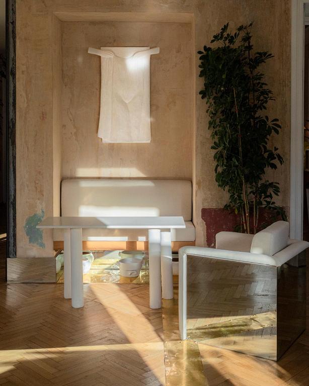 11 Unique Hotels in Georgia You Can't Miss! 8 Minimalist interior inspired by unique hotels in Georgia, with a beige wall, cream bench, white table and armchair, tall leafy plant, and sunlight casting warm patterns on the wooden floor. Sheer white fabric hangs framed above the bench.