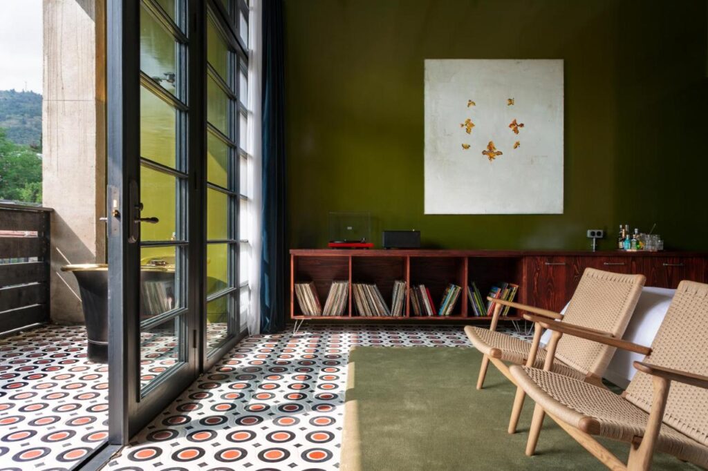 11 Unique Hotels in Georgia You Can't Miss! 5 A modern room in one of the unique hotels in Georgia, featuring green walls, a patterned tile floor, and sliding glass doors to a balcony. Two woven chairs face a wooden console with vinyl records, abstract art overhead, and natural light filling the space.