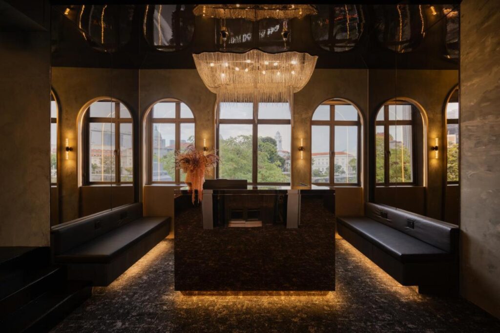 13 Most Unique Hotels in Singapore for an Incredible Holiday 20 A modern, dimly lit lounge in one of the unique hotels in Singapore, with black benches lining both sides, a sleek central bar, arched windows, and a large chandelier above, with greenery visible beyond the glass.