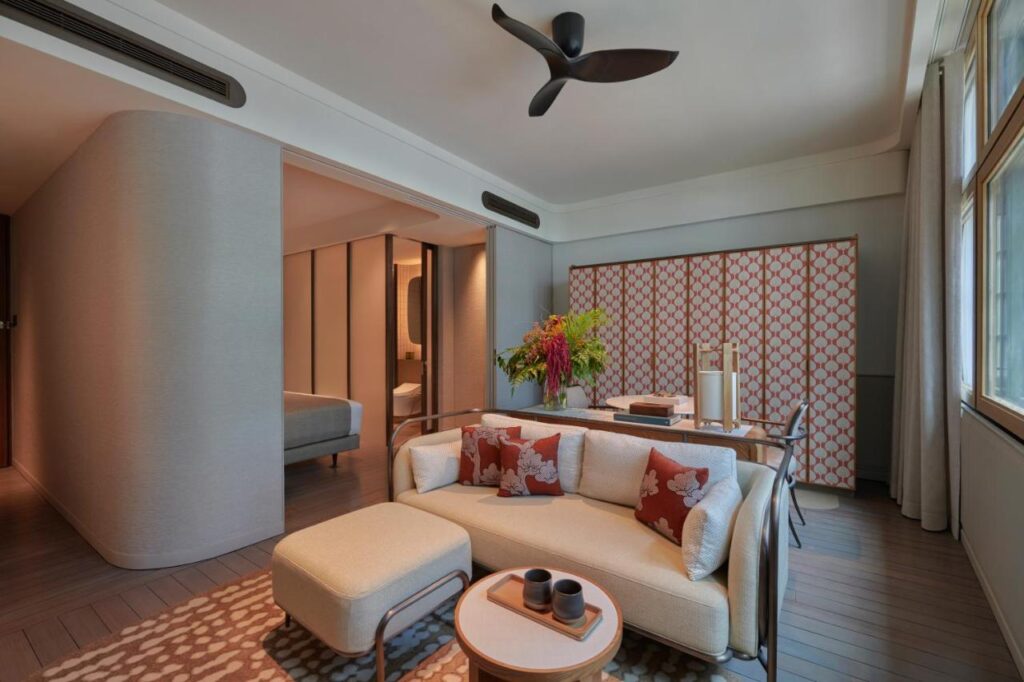 13 Most Unique Hotels in Singapore for an Incredible Holiday 35 A modern living room with a cream sofa, floral cushions, ottoman, round coffee table, and dining area—reflecting the stylish interiors often found in unique hotels in Singapore. Large windows, folding screens, and a glimpse of a bedroom complete the space.