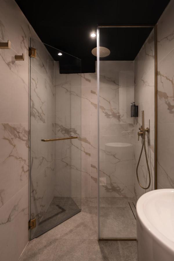 13 Most Unique Hotels in Singapore for an Incredible Holiday 23 A modern shower with glass doors, marble-patterned walls, a gold showerhead, and a black soap dispenser under a dark ceiling with recessed spotlights—reminiscent of the luxurious bathrooms found in unique hotels in Singapore.