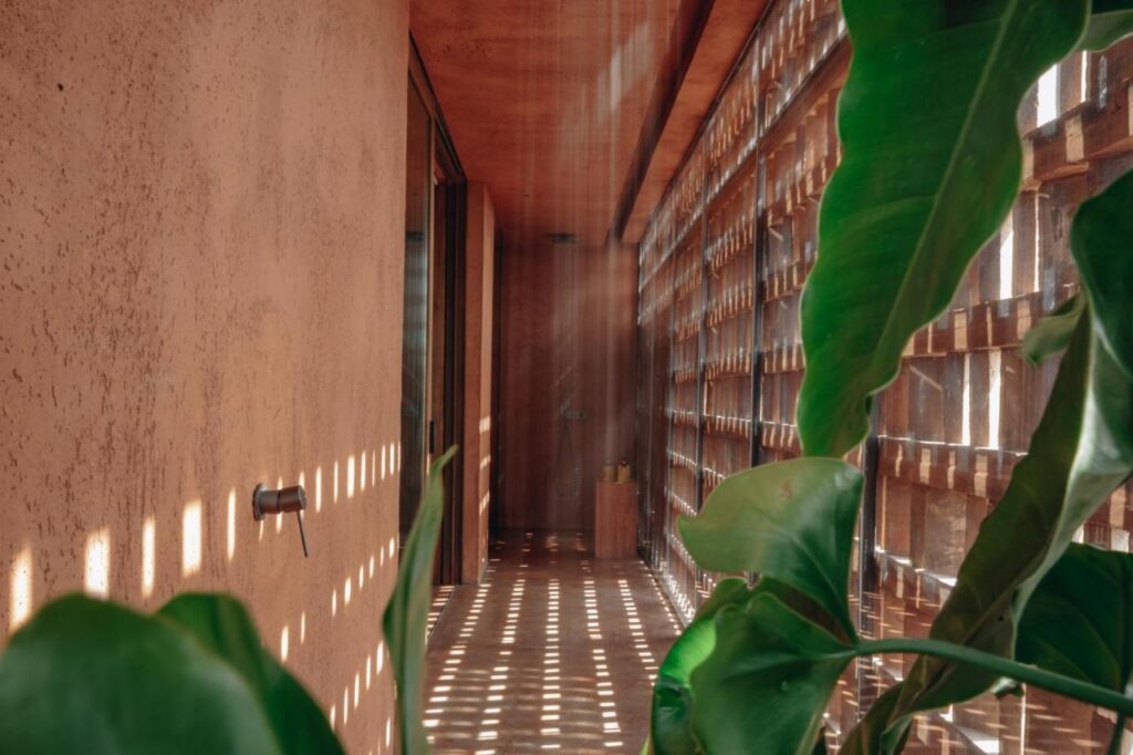 14 UNIQUE Boutique Hotels in Canggu, Bali 11 A narrow hallway with textured walls, sunlight streaming through a patterned screen creating light spots on the floor, and large green tropical leaves in the foreground—evoking the serene ambiance of boutique hotels in Canggu.