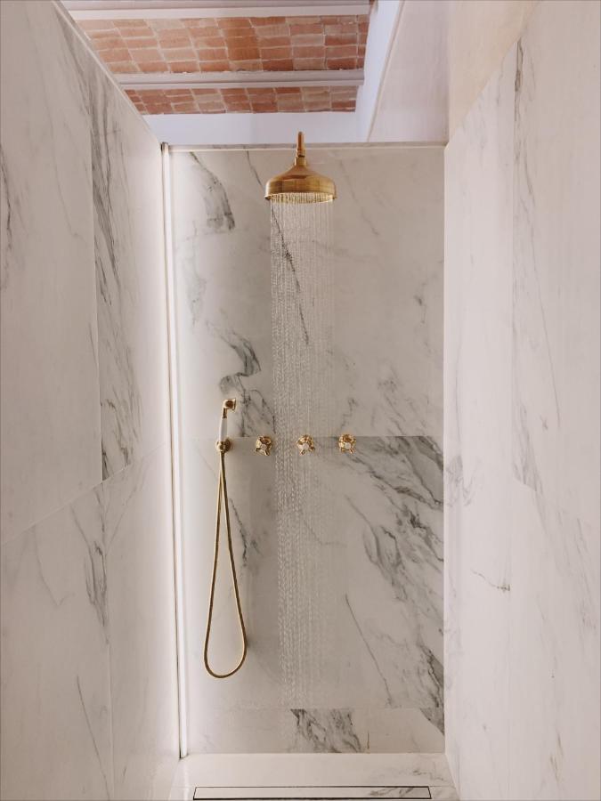 A modern shower with marble walls, a gold rainfall showerhead, matching gold fixtures, and a handheld attachment sits beneath an exposed brick ceiling—evoking the luxury found in boutique hotels in Costa Brava. Water cascades from the showerhead.