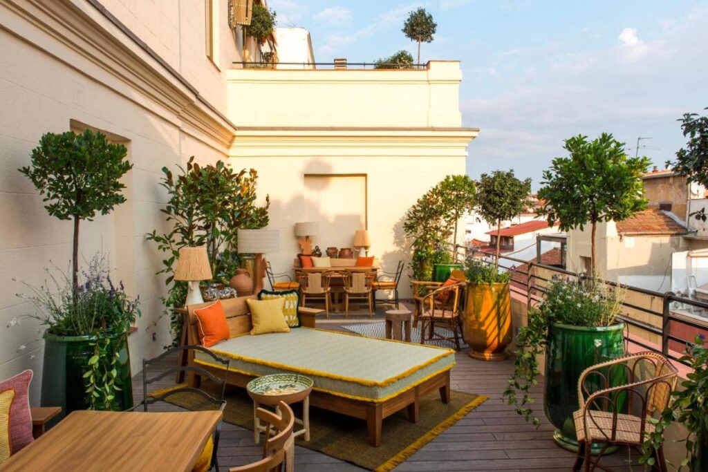 Where to Stay in Madrid: 14 Best Hotels 25 A stylish rooftop terrace with potted plants, a yellow-cushioned lounge chair, wicker chairs, and warm lighting—an inviting spot surrounded by greenery and rooftop views, perfect for those wondering where to stay in Madrid first time.