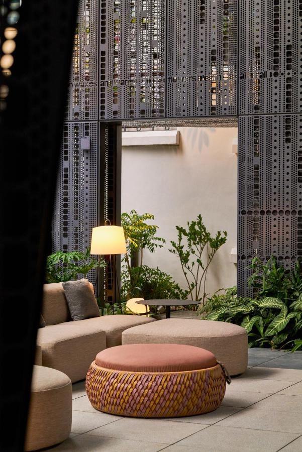 13 Most Unique Hotels in Singapore for an Incredible Holiday 34 Modern indoor lounge with beige sofas, a round woven ottoman, and floor lamp. Perforated metal wall panels, potted green plants, and natural light create the calm, contemporary atmosphere found in unique hotels in Singapore.