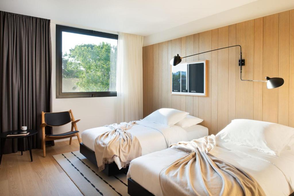 8 TOP Boutique Hotels in Split 24 A modern hotel room—perfect for those wondering where to stay in Split—features two single beds with white bedding, beige blankets, a large window with curtains, a wooden accent wall, black reading lamp, wooden chair, and small table.