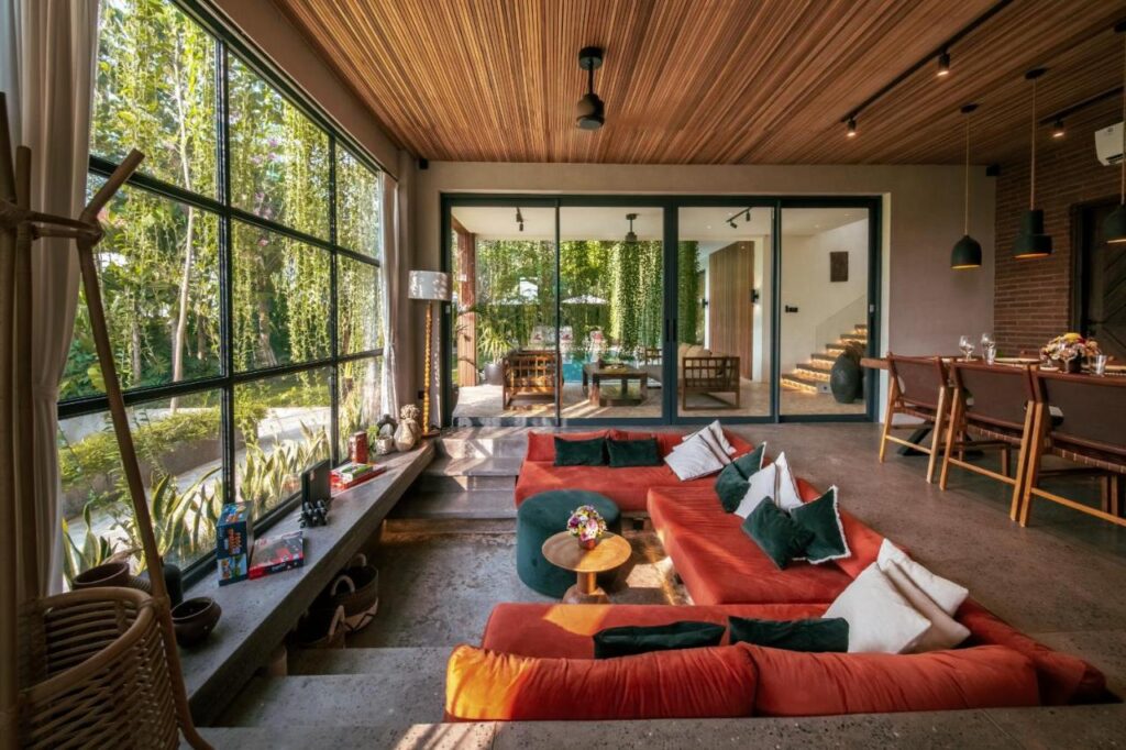 A modern living room in one of the best family villas in Bali, with large glass windows, sunken orange sofas, green and white pillows, a small round table, and garden views. Wooden ceiling panels and a stylish kitchen bar complete the space.