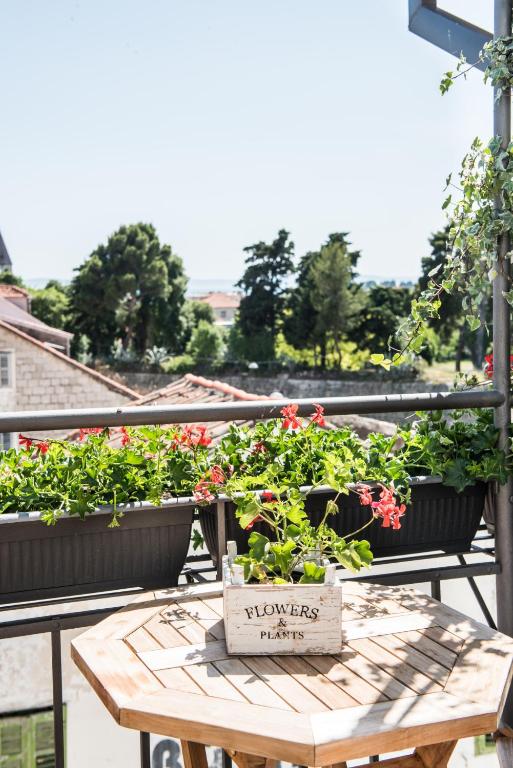 8 TOP Boutique Hotels in Split 14 A small wooden table with a flower box labeled FLOWERS & PLANTS sits on a balcony, surrounded by green plants and red flowers—an inviting spot for those seeking inspiration on where to stay in Split, overlooking trees and rooftops on a sunny day.