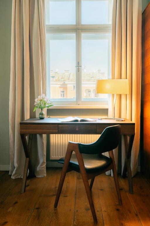 Unique Boutique Hotels in Salzburg 28 A wooden desk with an open book, a vase of flowers, and a lit lamp sits before a window with light curtains—evoking the charm of boutique hotels in Salzburg. A black chair rests by the desk on a gleaming wooden floor.
