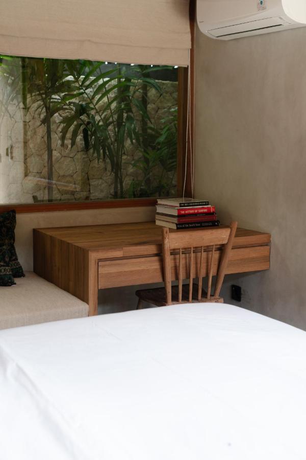 A small wooden desk with a stack of books and a wooden chair sits by a window overlooking greenery, evoking the cozy charm often found in the best family villas in Bali. Part of a bed with white bedding and a sofa are also visible in the inviting room.