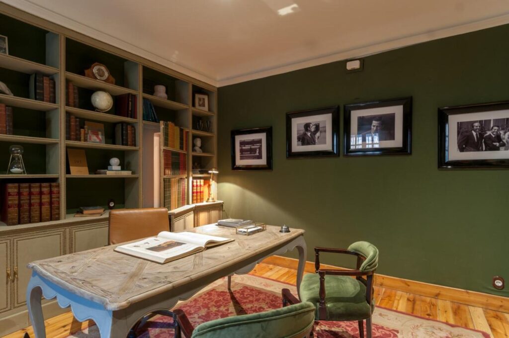 25 Weekend Trips from Madrid Max. 2h Away 100 A cozy home office with a wooden desk, green upholstered chairs, bookshelves filled with books and decor items—perfect for planning weekend trips from Madrid—framed black-and-white photos on an olive green wall, and a red patterned rug on a wooden floor.