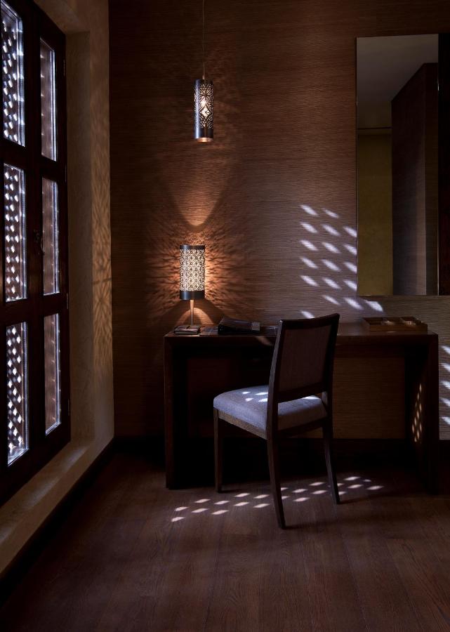 Where to Stay in Doha, Qatar: Transit, City & Beach Hotels 38 A stylish wooden desk and chair sit in a dimly lit room. Sunlight filters through patterned window screens, casting decorative shadows—a serene setting reminiscent of the peaceful interiors you might find when exploring where to stay in Doha.