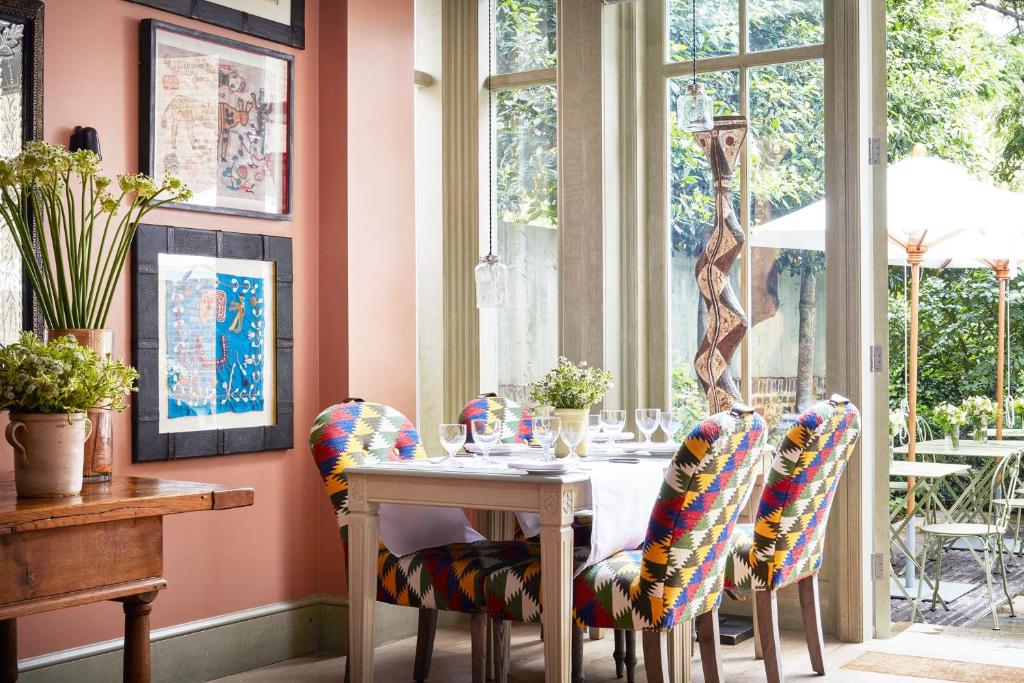15 Best Boutique Hotels in London, UK 57 A cozy dining area with colorful, patterned chairs around a set table by large windows, letting in natural light—reminiscent of boutique hotels in London. The walls are decorated with framed art, and greenery is visible outside.