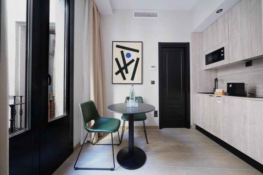 Where to Stay in Madrid: 14 Best Hotels 30 Modern kitchen and dining area with light wood cabinets, a round black table, two green chairs, a water bottle, and abstract framed art—perfect inspiration for those searching where to stay in Madrid first time. Large window with beige curtains lets in natural light.