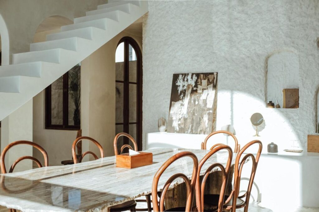 14 UNIQUE Boutique Hotels in Canggu, Bali 15 Bright, sunlit dining area with a marble table and eight wooden chairs. White textured walls, modern staircase, and art decor accents evoke the minimalist, airy style found in boutique hotels in Canggu.
