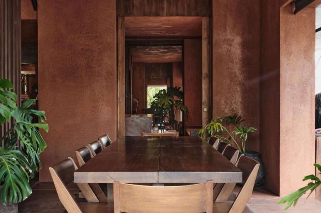 14 UNIQUE Boutique Hotels in Canggu, Bali 13 A wooden dining table with matching chairs is surrounded by green plants in a room with earthy brown walls, reminiscent of the modern, minimalist style often found in boutique hotels in Canggu. Natural light enters from the right, highlighting textures and decor.